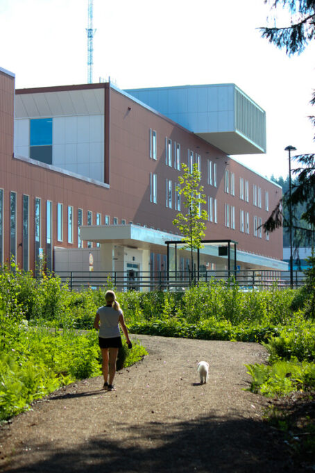 Nainen kävelee valkoisen pienen koiran kanssa kohti isoa modernia rakennusta, joka on Ratamokeskus eli terveysasema.