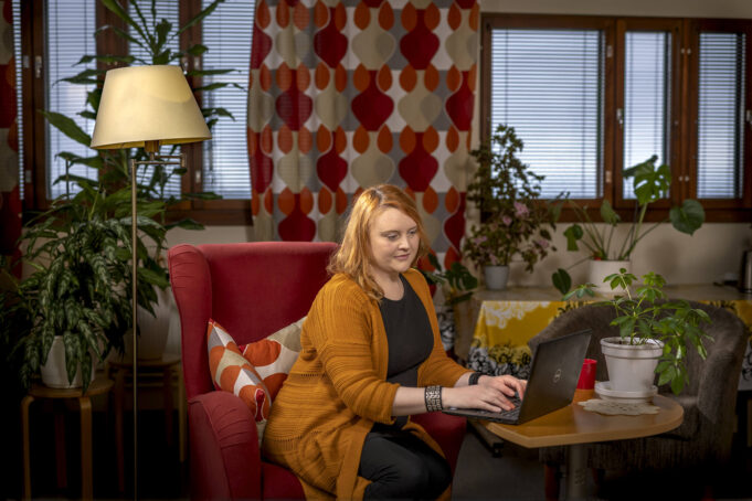 Henkilö punaisessa nojatuolissa näppäilee pöydällä olevaa kannettavaa tietokonetta.