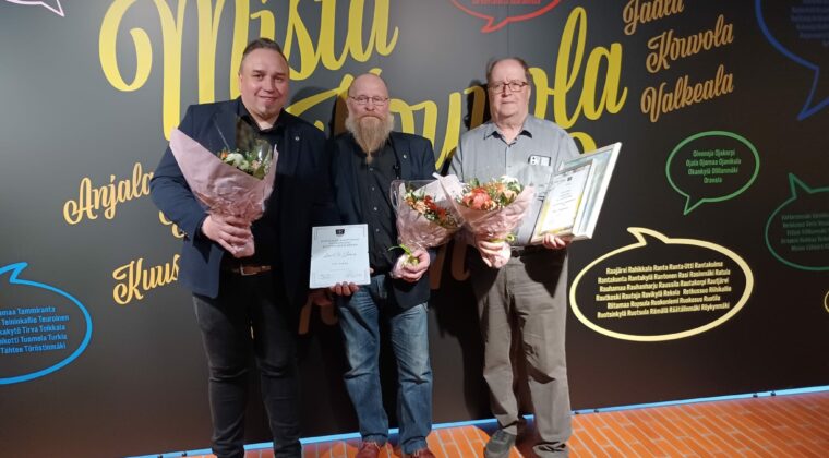 Kolme henkilöä seisoo palkintkukkien ja kunniakirjojen kanssa ja hymyilee kameralle.