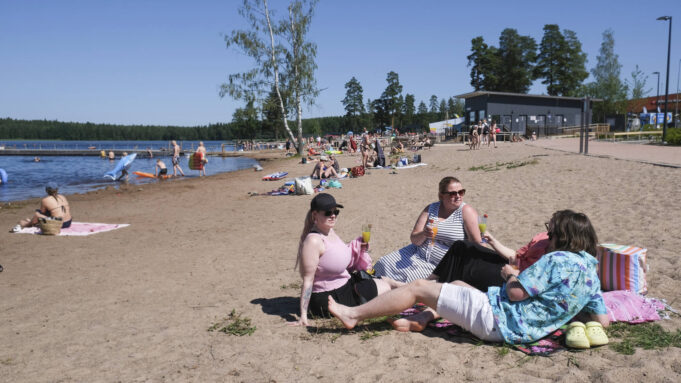 Ihmisiä uimarannalla keäsisissä asuissa. Taustalla järvi, laituri ja rakennuksia.