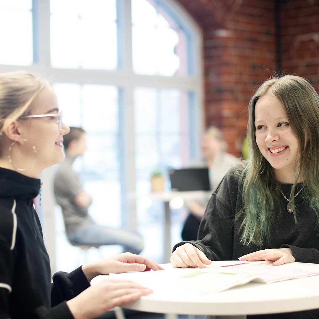 Kaksi hymyilevää nuorta naista pöydän ääressä. Tautsalla iso ikkuna ja tiiliseinää sekä muita ihmisiä.