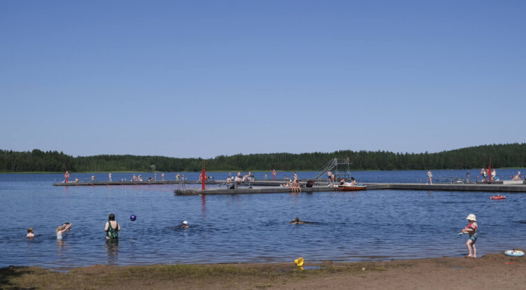 Uimareita järvessä. Edustalla hiekkaranta. Sininen pilvetön taivas.