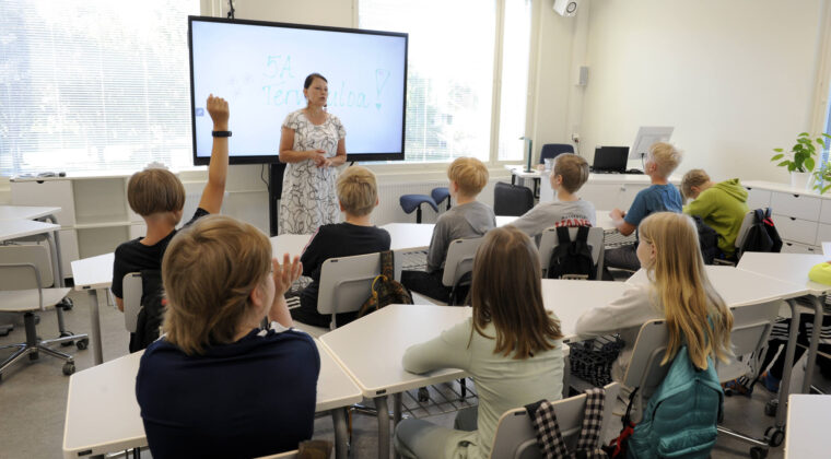 Oppilaita istuu pulpeteissa selkää kuvaajaan päin ja katsoo edessä olevaa opettajaa. Yksi oppilaista viittaa.