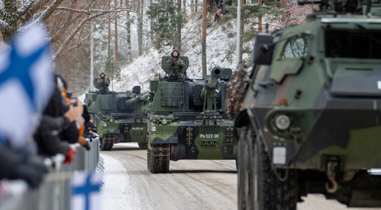Panssarivaunuja ajaa kadulla ja väkijoukko katselee vasemmalla tien laidassa. Suomen lippuja liehuu.