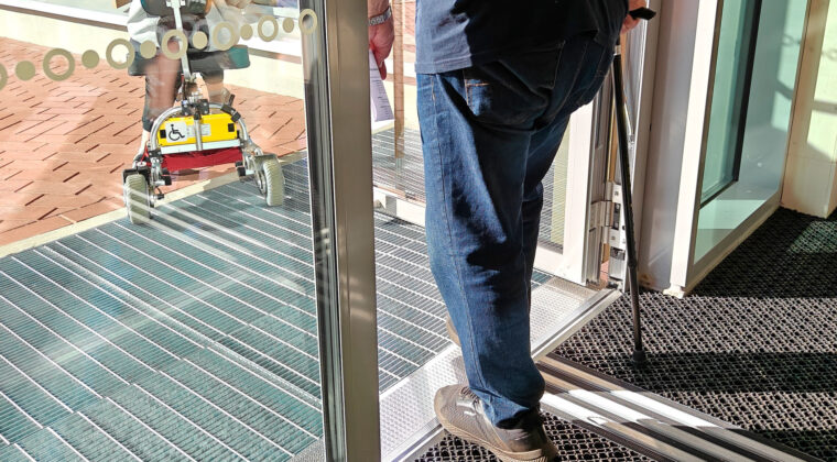 Oviaukosta pihalle kulkeehenkilö kepin kanssa. Pihalla näkyy henkilö, jolla on pyörillä kulkeva apuväline.