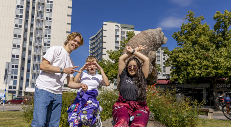 Kaksi opiskelijaa, joilla opiskelijahaalarit, istuvat karhupatsaan juurella. Yksi nuori seisoo vieressä.