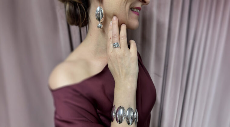 Henkilö viininpunaisessa ilta-asussa käsi leukapielessä. Ranteessa ja korvissa hopealusikoista tehdyt korut.