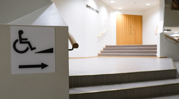 Kuvassa aulatila, jossa portaikkoja, seinässä merkki, jossa osoitetaan pyörätuolirampin olevan oikealla.