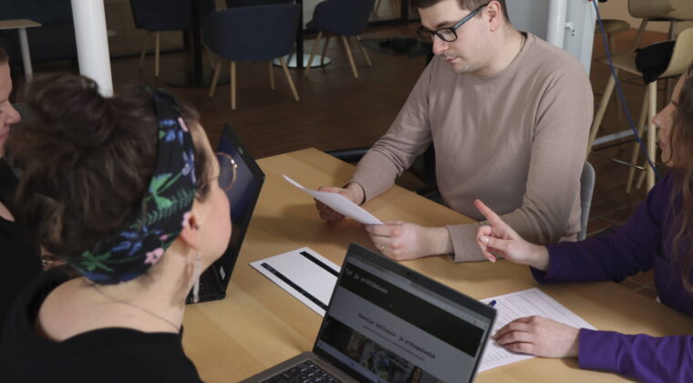 Henkilöitä keskustelemassa pöydän ääressä, pöydällä läppäri ja papereita.