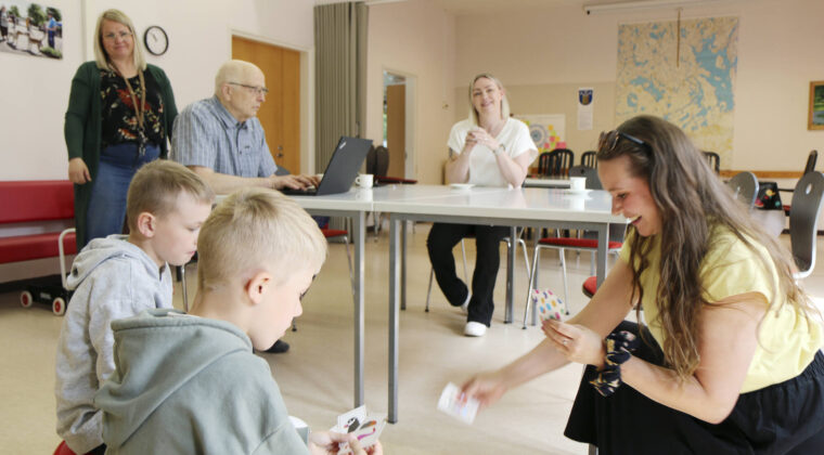 Aikuinen ja kaksi lasta pelaavat korttia sisätilassa. Taustalla muita ihmisiä pöydän ääreessä.