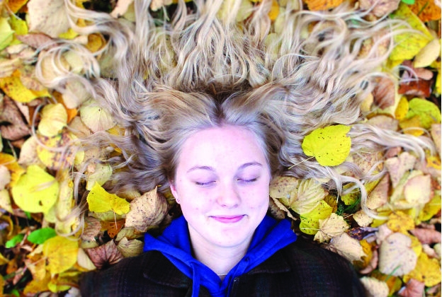 Pitkätukkainen henkilö makaa keltaisten lehtien päällä silmät kiinni ja hymyilee. Vaaleat pitkät hiukset ovat levälllään lehtien päällä.