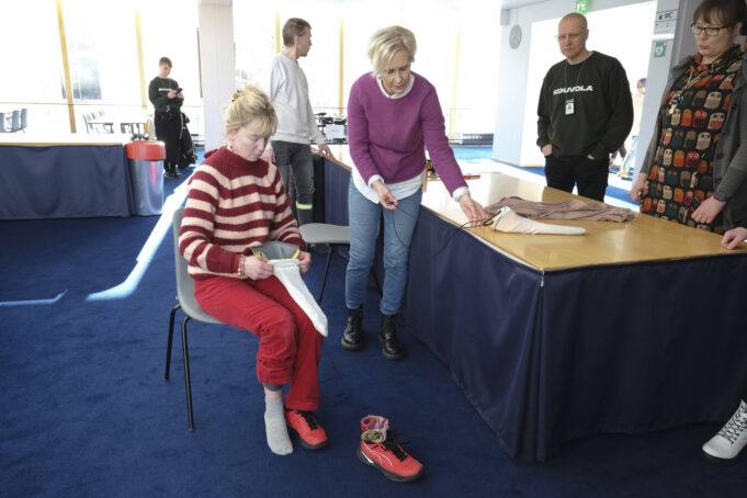 Nainen vetää sukkaa sukanpukija-apuvälineen päälle, toinen nainen näyttää mallia vieressä. Muutama henkilö seuraa toimitusta sivusta.