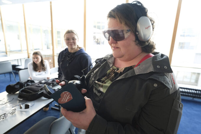 Nainen kuulosuojaimissa ja tummennetuissa laseissa käyttää puhelinta tekninen käsine kädessään. Taustalla kaksi nuorta henkilöä seuraa.