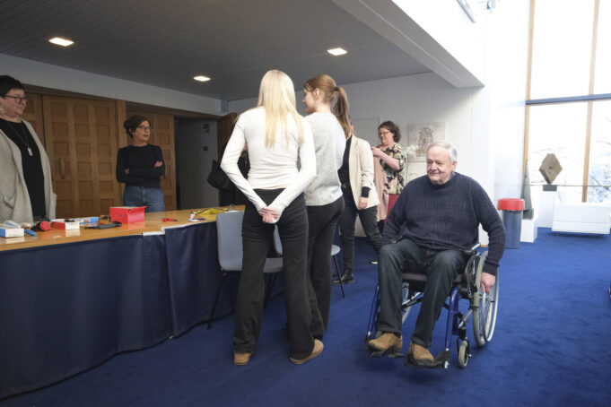 Mie kulkee pyörätuolilla tilaisuudessa, jossa ihmisryhmä keskustelee esittelypöydän ympärillä. Pöydällä on erilaisia apuvälineitä.