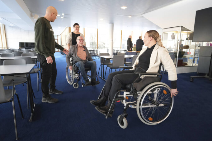 Ihmisjoukko Kouvolan kaupungintalon lämpiössä. Kaksi ihmistä istuu pyörätuoleissa ja yksi työntää toista pyörätuolia.