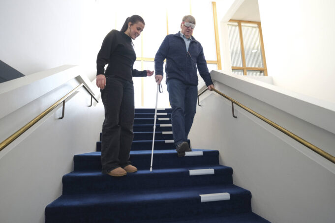 Mies peitetyissä silmälaseissa kulkee valkoinen keppi kädessään alas portaita. Vieressä nuori nainen vahtii toimitusta.