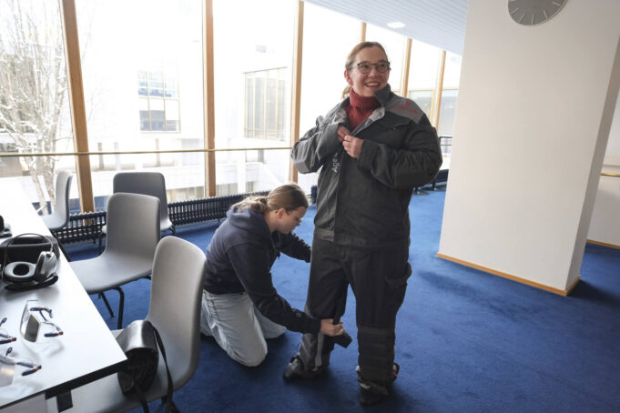 Nainen pukee työasun näköistä asua päälleen. Nuori nainen sitoo jalkatukea hänen toiseen jalkaansa.