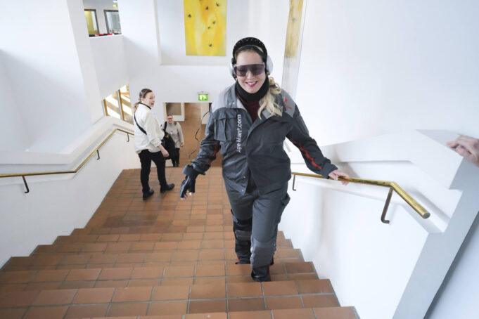 Nainen työasu (ikäpuku) päällään, kuulosuojaimet ja suojalasit päässään nousee rappuja. Hän pitää toisella kädellä kiinni kaiteesta ja toisessa on musta, tekninen kansikas.