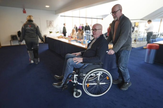 Mies työntää toista pyörätuolissa salissa, jossa on esittelypöytä ja useampia ihmisiä tilassa keskustelemassa keskenään.