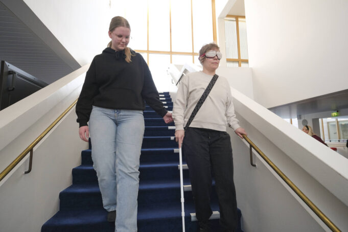 Nainen ohjaa kädellä toista naista, jonka silmät on peitetty ja jolla on valkoinen keppi kädessään.