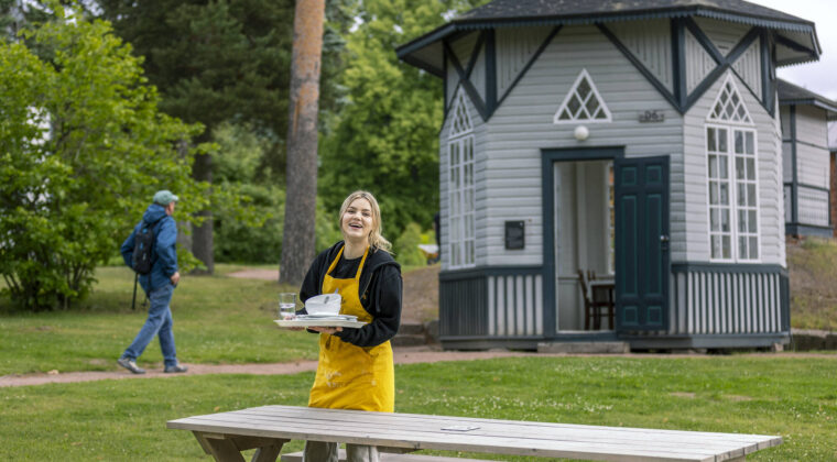 Keltaessuinen henkilö kantaa lautasia ja hymyilee kameralle.