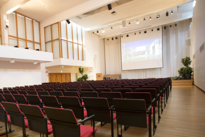 A large hall with light-coloured walls, rows of red seats, and a stage with a projection screen showing a slide presentation.
