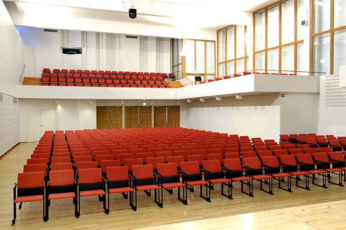 A hall with light-coloured walls and rows of red seats. At the back of the hall there is a balcony with additional rows of seats.