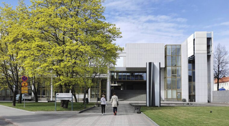 Vaalea, suuri julkinen rakennus, jossa suuret raput nousevat rakennuksen sisustaa kohti. Kaksi henkilöä kulkee kohti rakennusta. Kouvolan kaupungintalo.