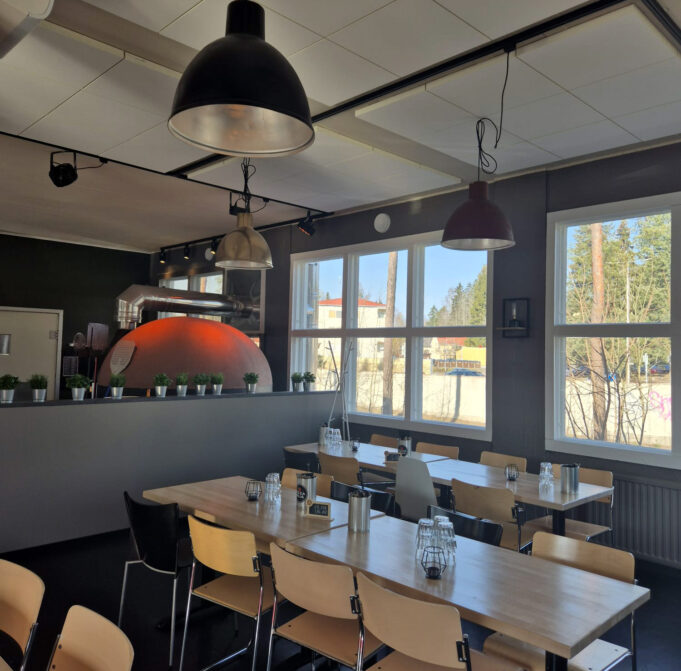 A restaurant with a pizza owen. The room has big windows and table groups.