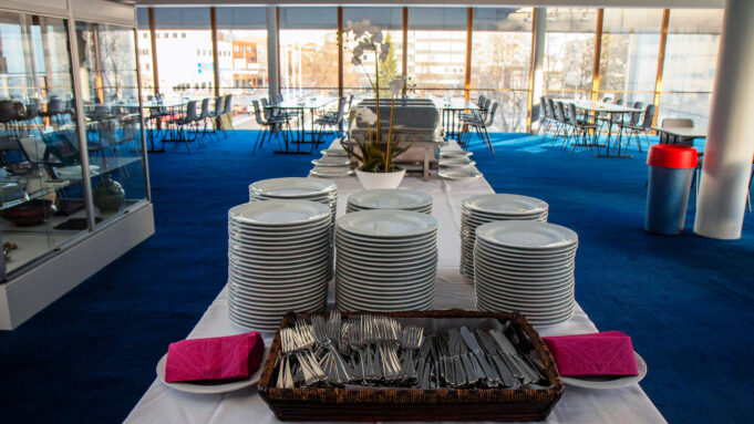 A serving table with cutlery in a spacious room, the wall of which is filled with large windows overlooking the city.