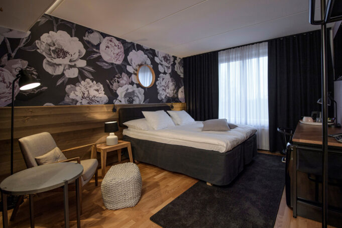 Hotel room with a wide bed, dark floral feature wall and wood panelling, a small seating area with chairs and table, and a desk beside a large window with dark curtains.
