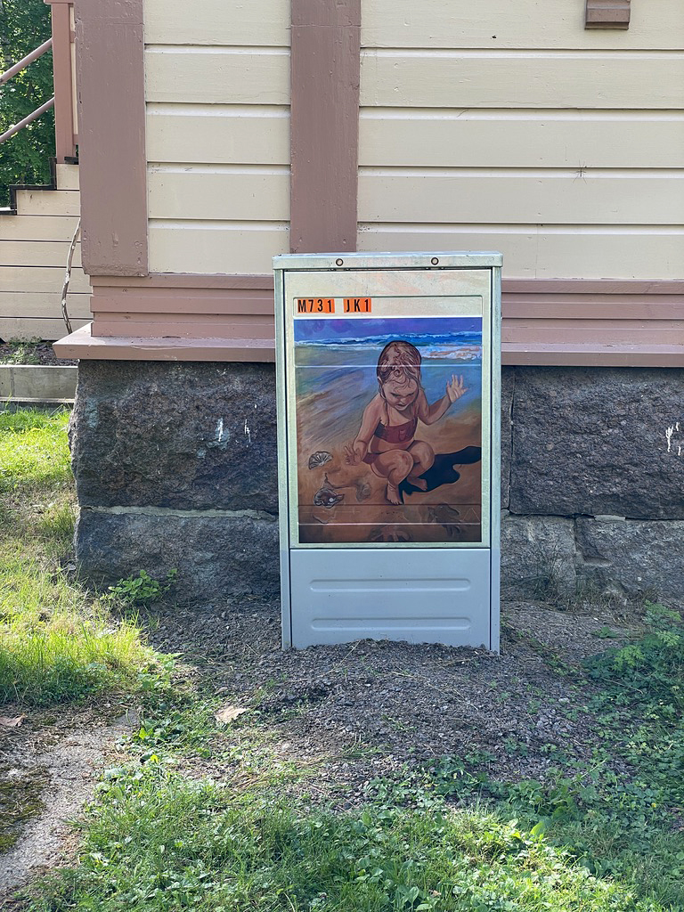 Maalattu sähkökaappi vanhan puurakennuksen juurella. Teoksessa lapsi leikkii uimapuvussa uimarannalla.