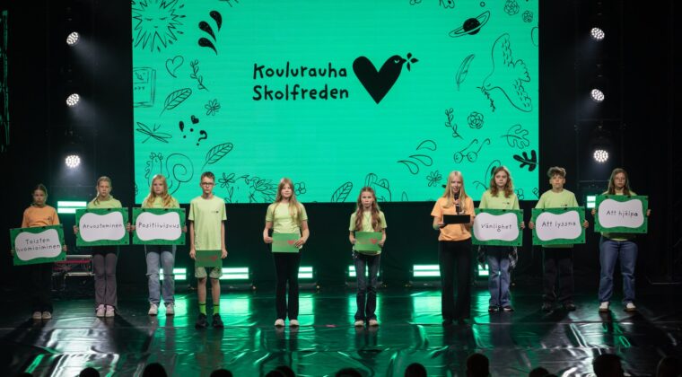 Oppilaita rivissä lavalla erilaisia kylttejä kädessään. Taustalla vihreä kuva, jossa lukee "koulurauha - skolfred".