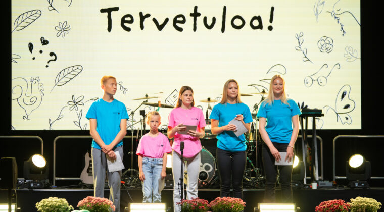 Viisi henkilöä erivärisissä t-paidoissa seisoo lavalla, jonka takana seinällä lukee "Tervetuloa!".