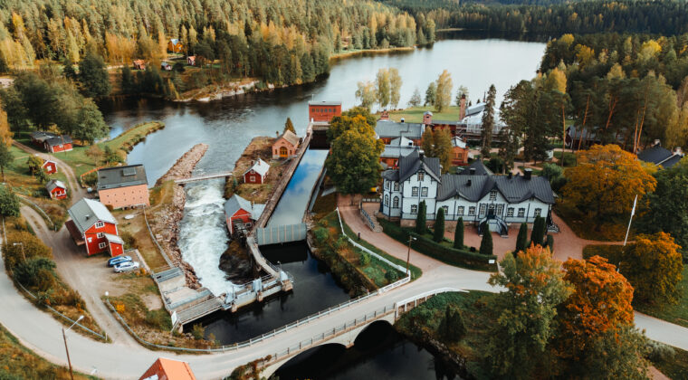 Ilmakuva syksyisestä Verlan alueesta jossa näkyy rakennuksia ja joki sekä joen yli kulkeva silta.
